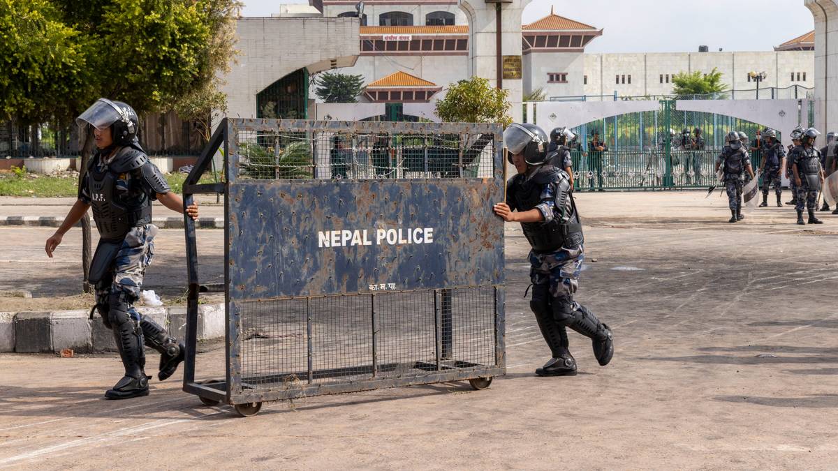 Funkcjonariusze policji w pełnym rynsztunku, w tym kaskach ochronnych i kamizelkach kuloodpornych, pchają metalową blokadę drogową z napisem "NEPAL POLICE".