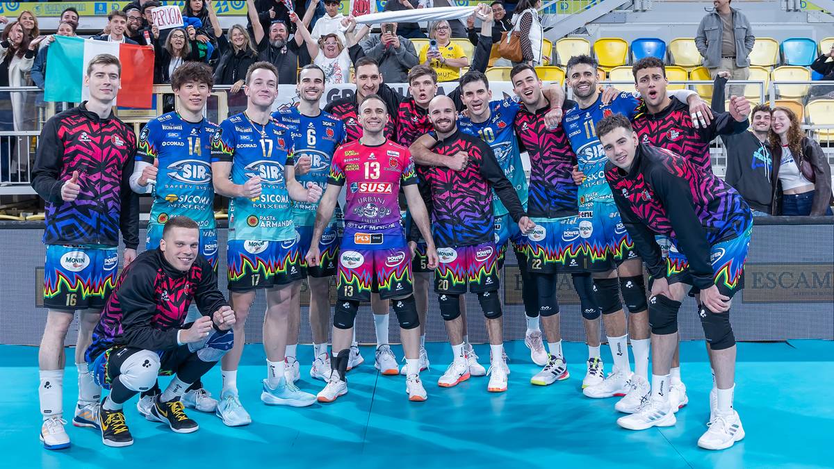 Drużyna siatkarzy pozuje do zdjęcia na parkiecie hali sportowej, ubrana w niebieskie i czerwono-zielone stroje.