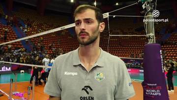 Mężczyzna z kr&oacute;tkimi ciemnymi włosami i brodą stoi na boisku do siatk&oacute;wki, ubrany w szarą koszulkę polo z logo "Kempa" i "Orlen". W tle widać trybuny wypełnione kibicami oraz siatkę do gry.