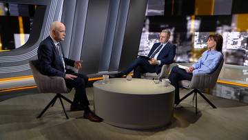 Trzy osoby siedzą w studiu telewizyjnym przy okrągłym stoliku. Dw&oacute;ch mężczyzn i jedna kobieta biorą udział w dyskusji. Na stole znajdują się kubki. W tle widać ekran z grafikami.