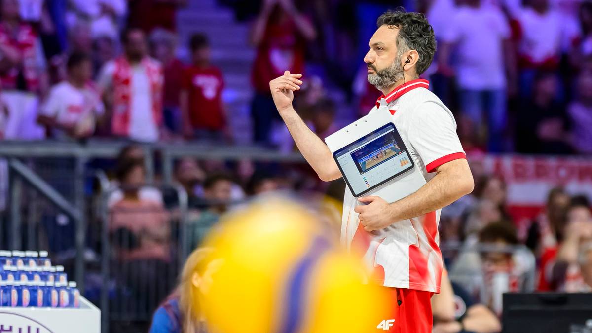 Trener siatk&oacute;wki w białej koszulce z czerwonymi elementami instruuje zawodnik&oacute;w, trzymając tablet.