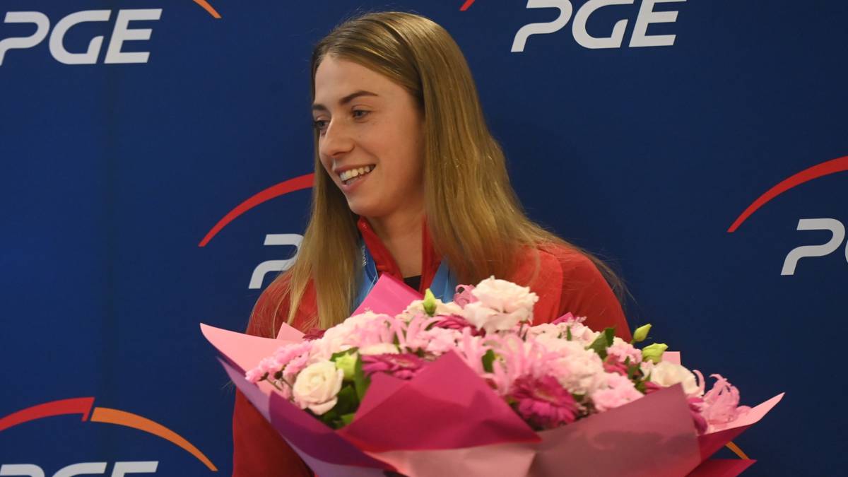 Uśmiechnięta młoda kobieta z blond włosami, ubrana w czerwoną bluzę, trzyma bukiet różowych i białych kwiatów.