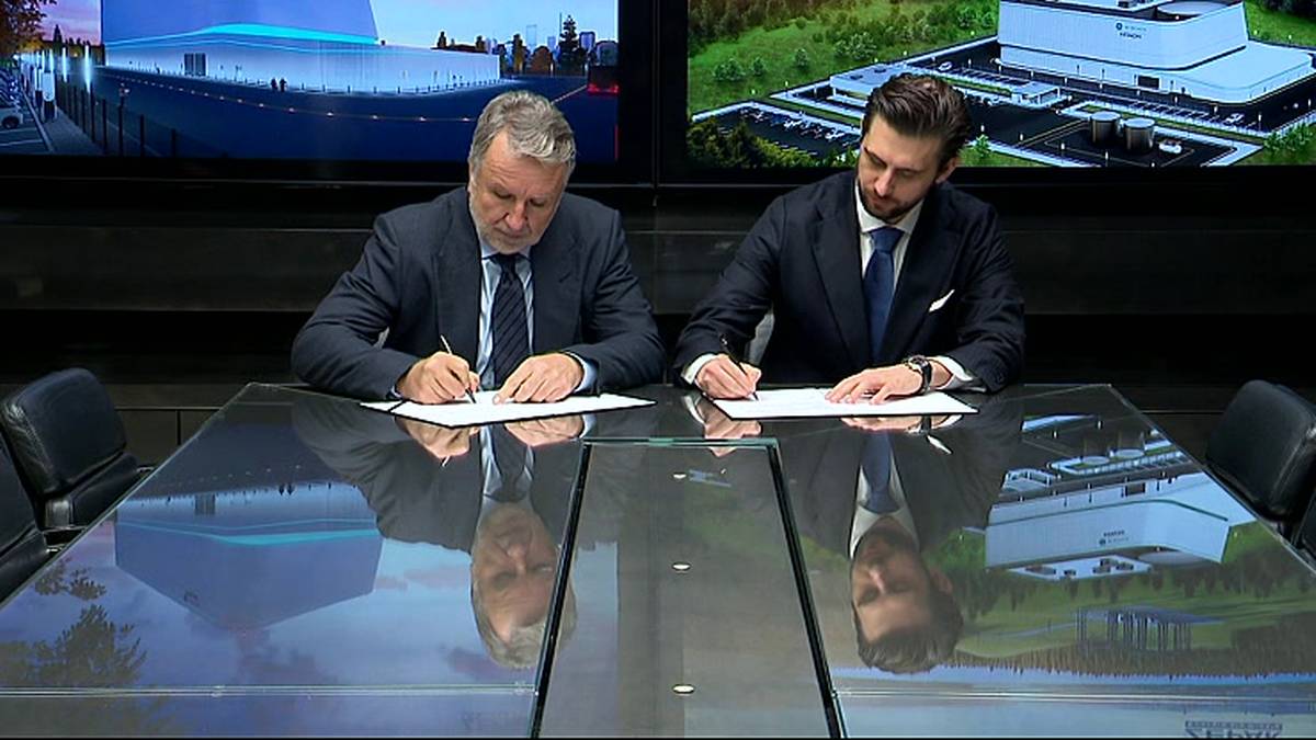 Dw&oacute;ch mężczyzn w garniturach podpisuje dokumenty przy szklanym stole na tle wizualizacji budynk&oacute;w.