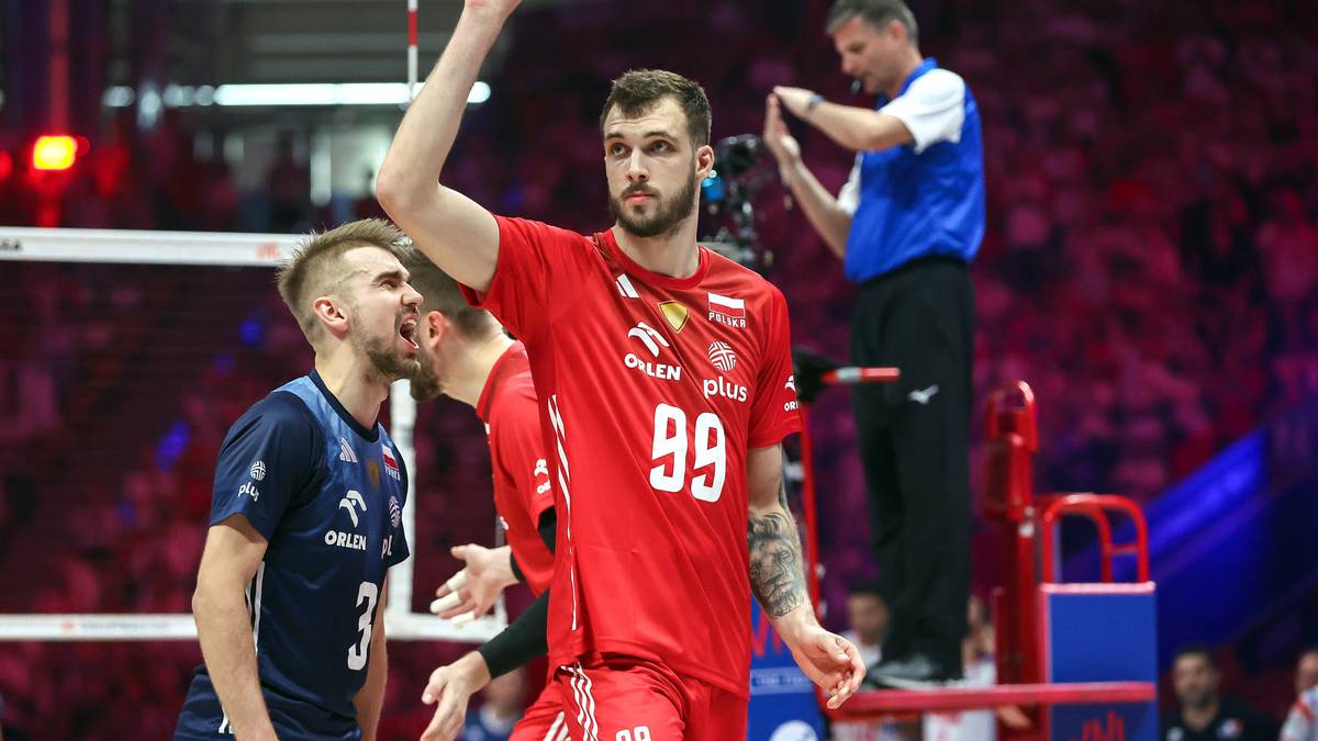 Siatkarz w czerwonej koszulce z numerem 99 na plecach, podnosi rękę w geście triumfu. Obok niego stoi inny siatkarz w niebieskiej koszulce z numerem 3. W tle widać fragment boiska i trybuny wypełnione kibicami.