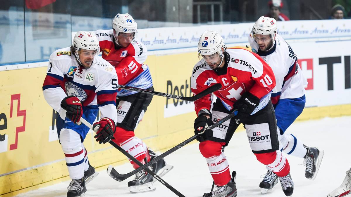 Czterech zawodników hokejowych w akcji na lodowisku, walczących o krążek. Dwóch w czerwonych strojach, dwóch w białych.