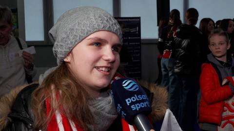 Młoda dziewczynka w szarej czapce i szaliku z białymi i czerwonymi pasami uśmiecha się, trzymając mikrofon "Polsat Sport" podczas wywiadu. W tle widoczni są inni ludzie.