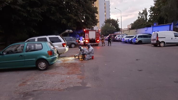 Na osiedlowym parkingu, przy zaparkowanych samochodach, dwie osoby w kombinezonach ochronnych pracują nad usunięciem substancji przypominającej rtęć, która rozlała się na ziemi. W tle widoczny jest samochód strażacki z włączonymi sygnałami świetlnymi oraz budynek mieszkalny.