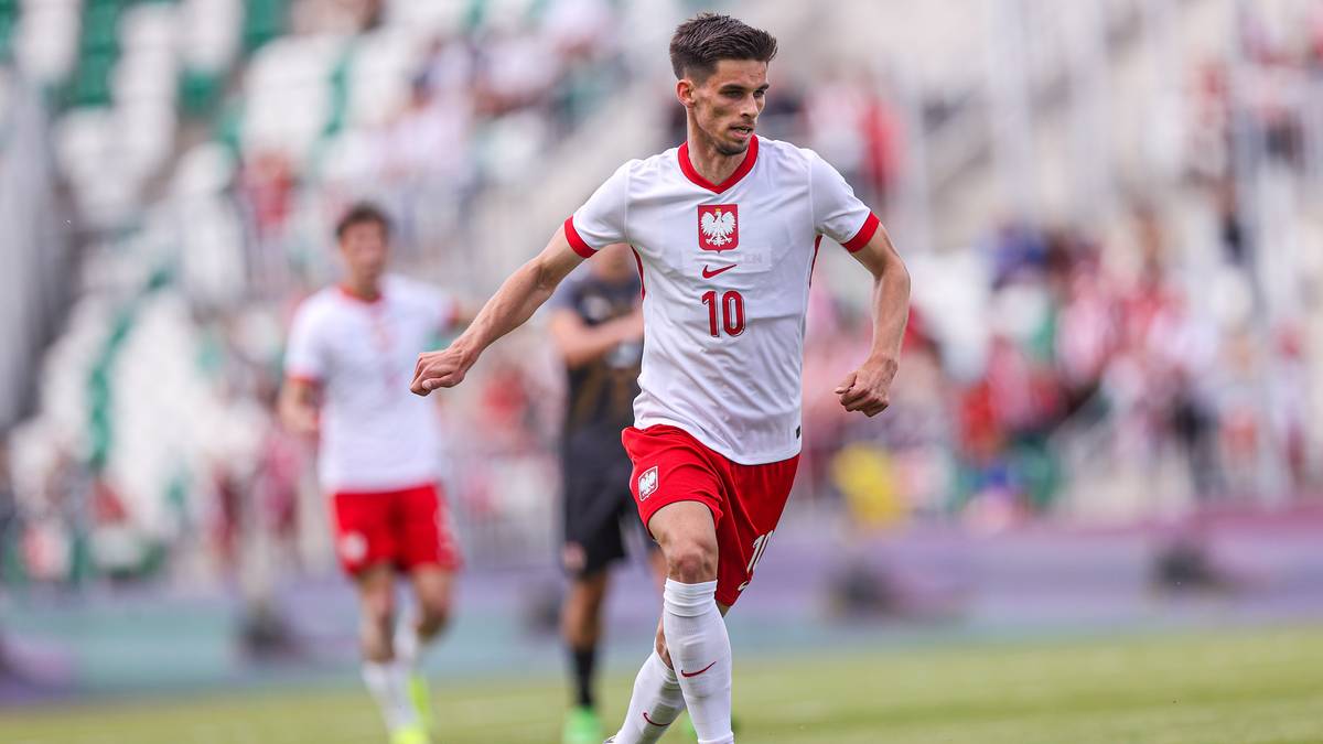 Piłkarz w białej koszulce z numerem 10 i czerwonych spodenkach prowadzi piłkę na boisku.