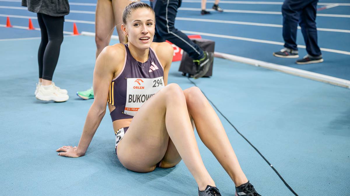 Lekkoatletka siedząca na niebieskiej bieżni, ubrana w sportowy strój, z numerem startowym 294 i nazwiskiem BUKOWIECKA. Wygląda na zmęczoną po zawodach.