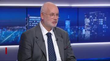 Starszy mężczyzna w okularach i garniturze, z krawatem w granatowo-białe wzory, siedzi przed ekranem z widokiem na nocne miasto.