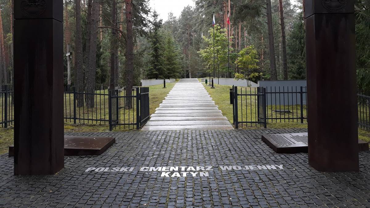 Wejście na Polski Cmentarz Wojenny w Katyniu z dwoma ciemnymi obeliskami po bokach i kamienną alejką prowadzącą w głąb lasu.
