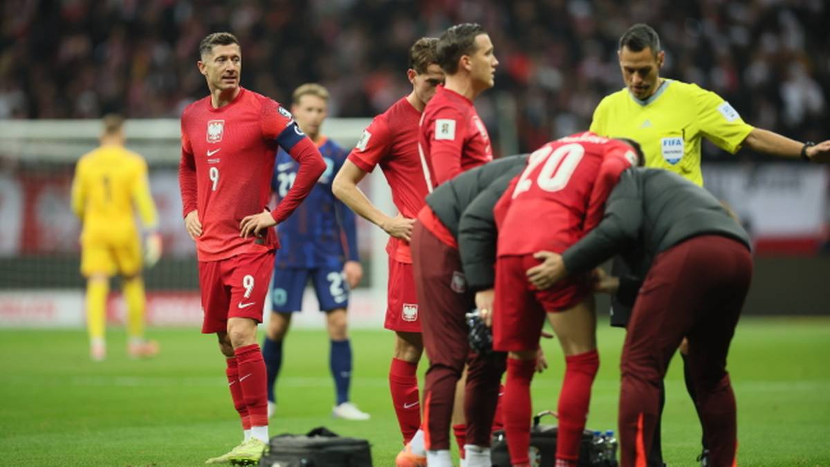 Piłkarze w czerwonych strojach otaczają kontuzjowanego kolegę na boisku piłkarskim, w tle sędzia.