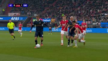 Mecz piłki nożnej pomiędzy AZ Alkmaar a Go Ahead Eagles. Gracze w niebieskich i czerwonych strojach rywalizują o piłkę na zielonej murawie stadionu, na tle trybun wypełnionych kibicami. Wynik meczu wyświetlany w górnym rogu ekranu: AZ 0 - 1 GAE.