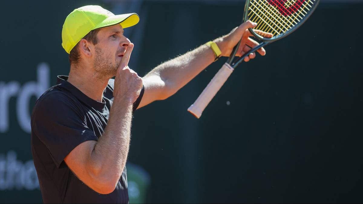 Tenisista Hubert Hurkacz w żółtej czapce, czarnej koszulce i z rakietą w ręku, z palcem wskazującym na ustach, wykonuje gest ciszy.