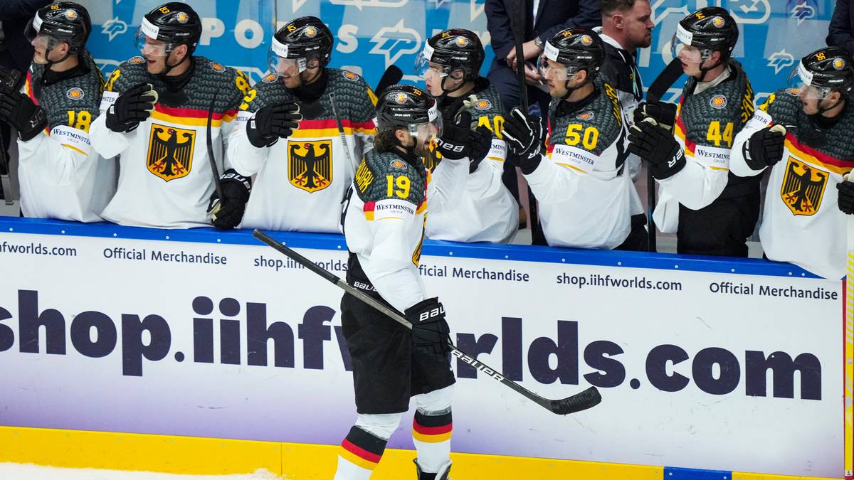 Hokeista w stroju reprezentacji Niemiec z numerem 19 na plecach stoi na lodowisku z kijem w dłoni, za nim widać ławkę rezerwowych z innymi zawodnikami w identycznych strojach.