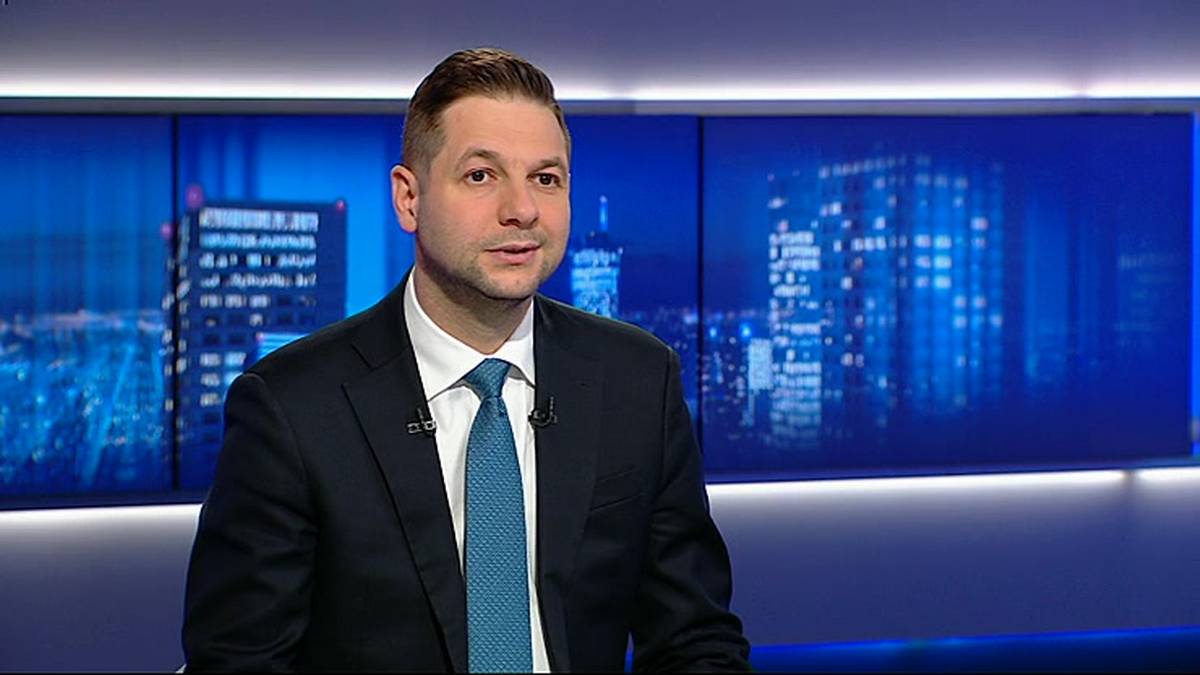 Mężczyzna w garniturze i krawacie siedzi na tle ekran&oacute;w z widokiem nocnego miasta.