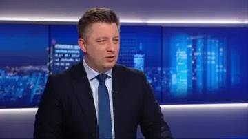 Michał Dworczyk w garniturze i krawacie, wypowiadający się przed niebieskim tłem z widokiem na nocne miasto.