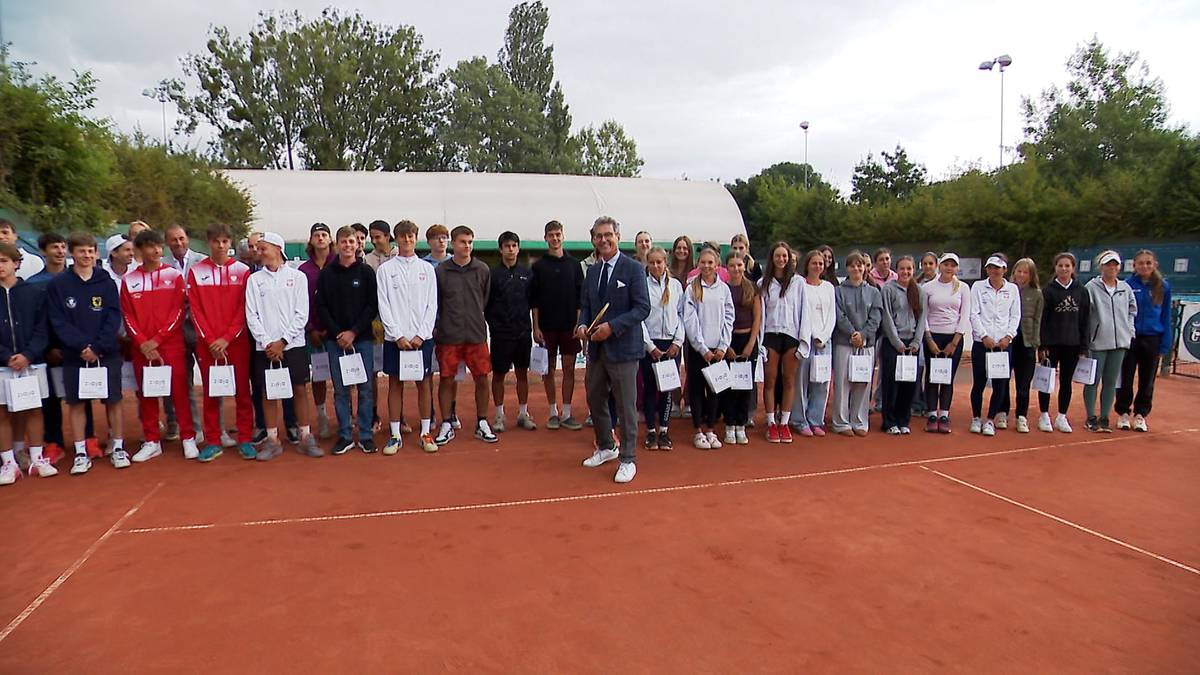 Grupa osób, głównie młodych sportowców, stoi w rzędzie na korcie tenisowym. W środku stoi mężczyzna w garniturze, prawdopodobnie organizator lub gość honorowy. Wszyscy uczestnicy trzymają w rękach białe koperty. W tle widoczne są białe namioty i zielona roślinność.