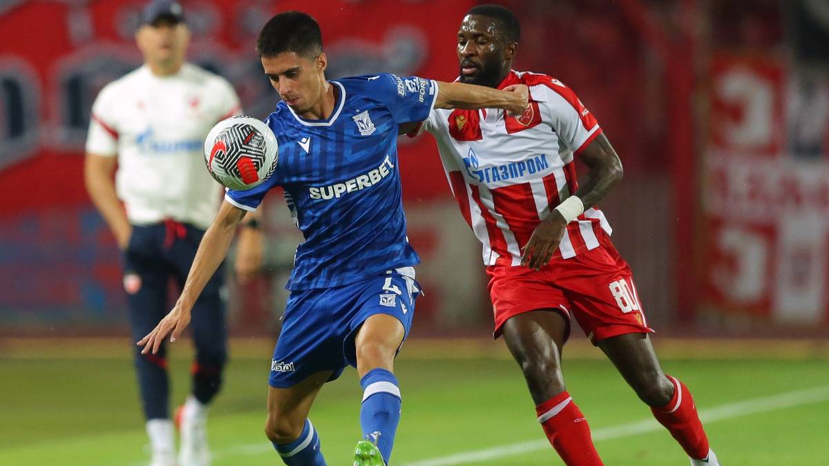 Piłkarz w niebieskiej koszulce Lecha Poznań drybluje z piłką, podczas gdy drugi piłkarz w czerwono-białej koszulce próbuje go zatrzymać.