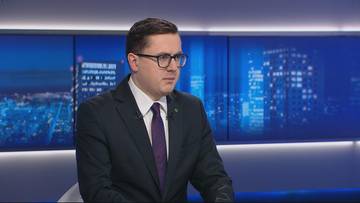 Mężczyzna w garniturze i okularach siedzi przed ekranem z widokiem nocnego miasta.