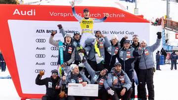 Grupa narciarzy na podium z czekiem i pucharami, w tle tablica wynik&oacute;w zawod&oacute;w Audi Ski World Cup.