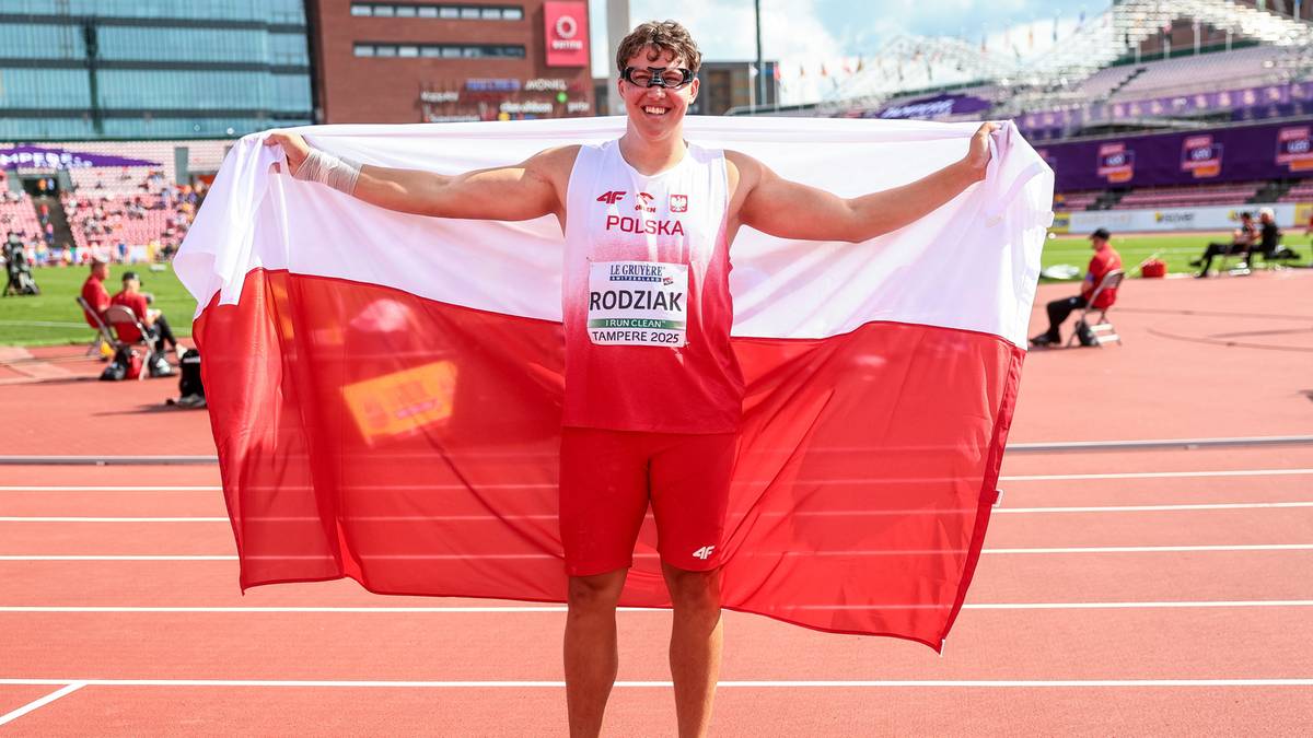 Atletycznie zbudowany mężczyzna w stroju sportowym z napisem "POLSKA" i nazwiskiem "RODZIAK" na piersiach, stoi na bieżni stadionu lekkoatletycznego, trzymając rozłożoną biało-czerwoną flagę Polski za sobą.