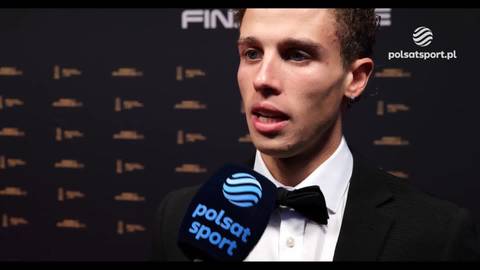 Mężczyzna w czarnym garniturze i muszce z mikrofonem "polsat sport" przed nim, mówiący do kamery.