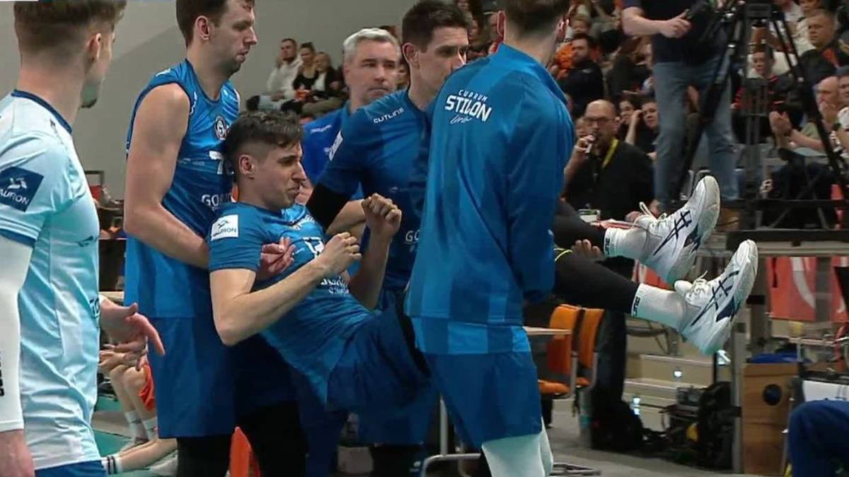 Siatkarz w niebieskiej koszulce trzyma za rękę innego zawodnika w tej samej drużynie, kt&oacute;ry wygląda na zranionego i siedzi na parkiecie.