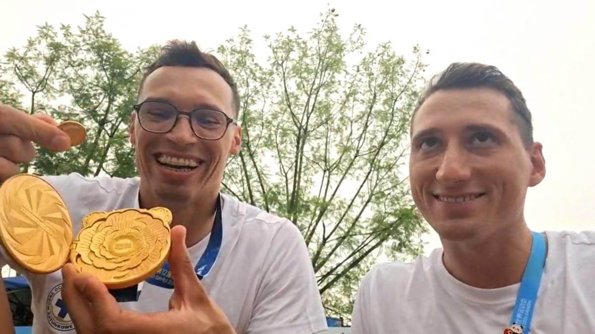 Dwóch uśmiechniętych młodych mężczyzn w koszulkach, trzymających złote medale. Jeden z nich, po lewej, pokazuje dwa medale do kamery. Na szyjach obu mężczyzn widoczne są niebieskie szarfy. W tle widać zielone drzewa i niebo.