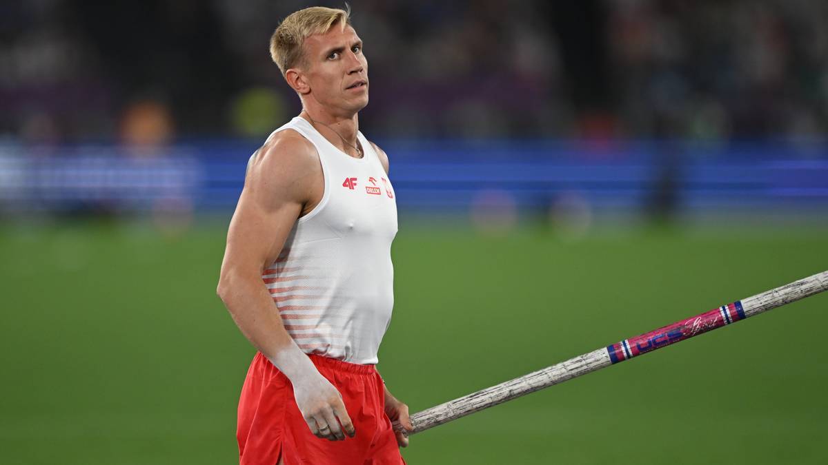 Atletka w białe koszulce i czerwonych spodenkach trzyma tyczkę, patrząc w g&oacute;rę.