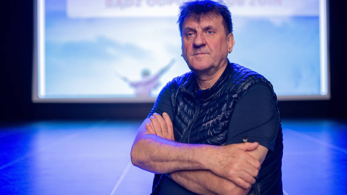 Marek Kamiński stoi z rękami skrzyżowanymi na piersi, na tle ekranu z grafiką przedstawiającą człowieka na tle nieba.