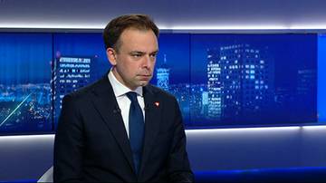 Mężczyzna w ciemnym garniturze i granatowym krawacie siedzi przed niebieskimi ekranami, na kt&oacute;rych wyświetlone są nocne panoramy miast z wieżowcami.