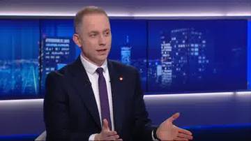 Mężczyzna w garniturze i krawacie m&oacute;wi, gestykulując rękami. W tle widać ekran z widokiem nocnego miasta.