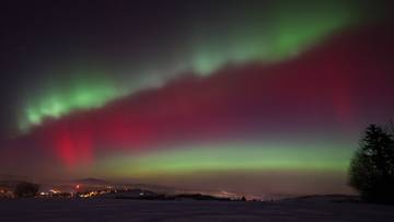 Widok na nocne niebo z zorzą polarną w kolorach zielonym, czerwonym i purpurowym, nad krajobrazem z widocznymi światłami miasta i drzewami.