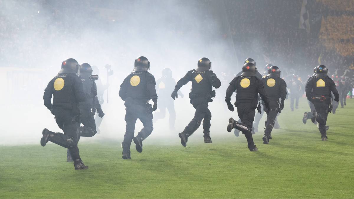 Grupa policjant&oacute;w w pełnym rynsztunku biegnie po murawie stadionu, w tle unosi się dym.