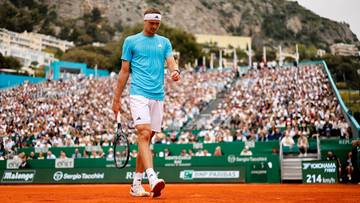 Tenisista w niebieskiej koszulce i białych spodenkach idzie po korcie ziemnym, trzymając rakietę w prawej ręce. W tle widać trybuny wypełnione widzami i budynki.