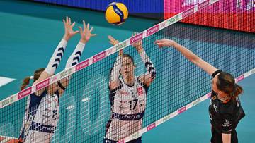 Siatkarki podczas meczu pr&oacute;bują zablokować atak przeciwniczki nad siatką.