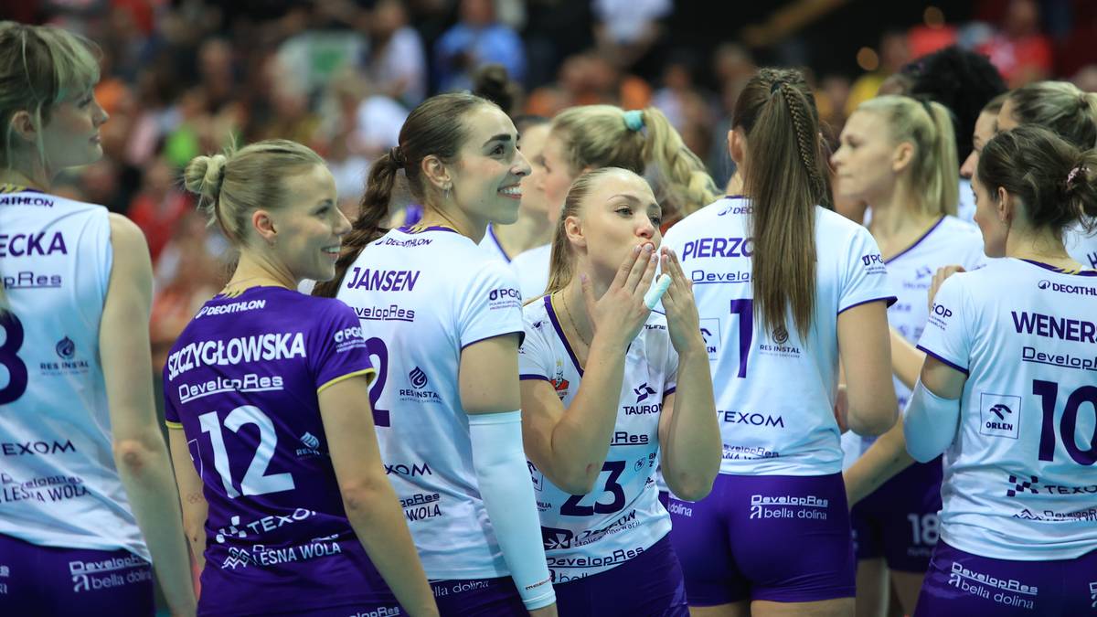 Grupa siatkarek w strojach sportowych, część w białych, część w fioletowych.