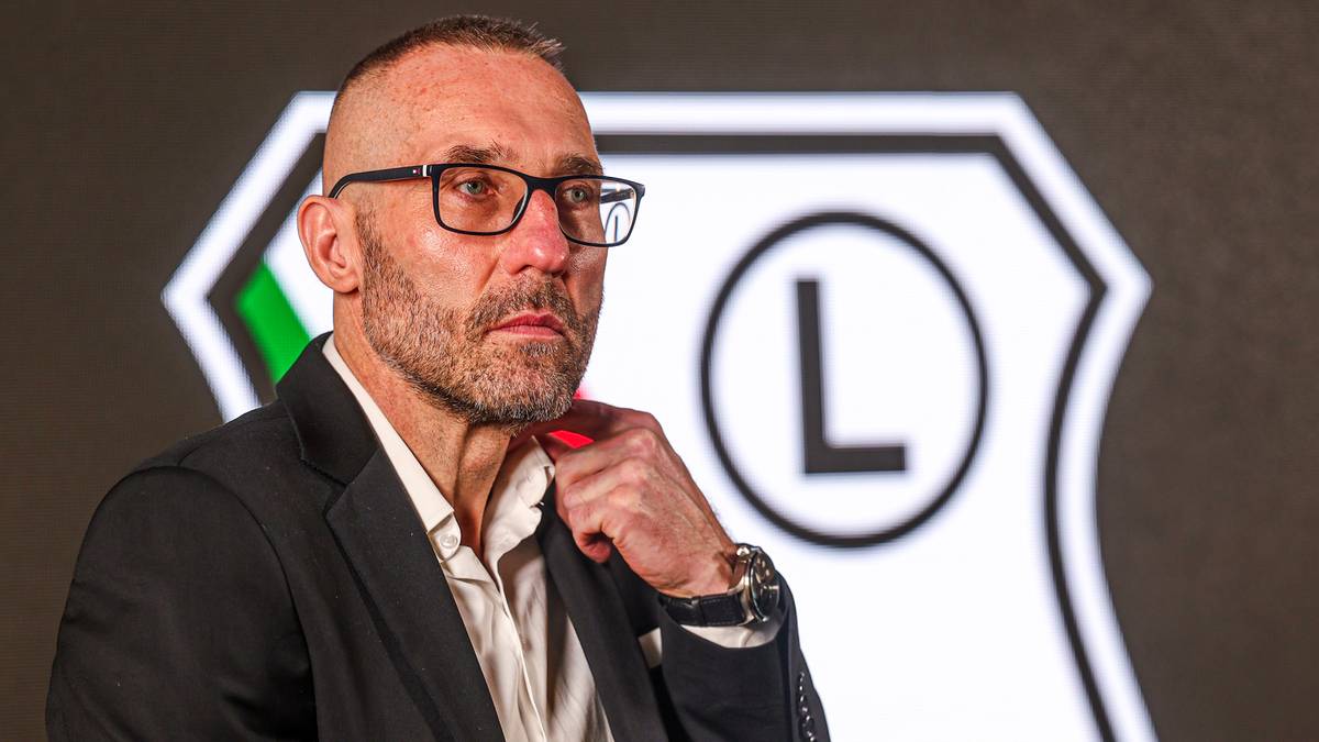 Mężczyzna w okularach i garniturze z zarostem, na tle logo klubu sportowego.