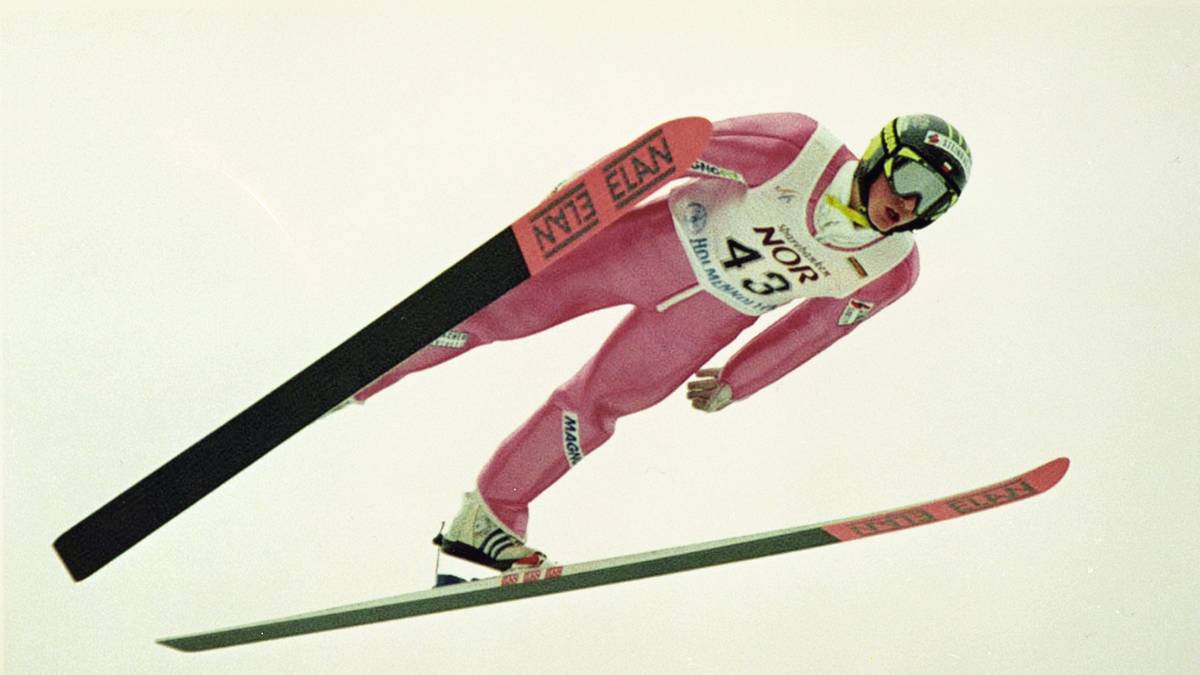 Skoczek narciarski w r&oacute;żowym kombinezonie i kasku, wykonujący skok na nartach.