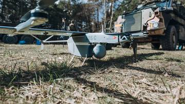 Dron z widocznymi śmigłami i konstrukcją, ustawiony na trawie. W tle znajduje się pojazd wojskowy i drzewa.