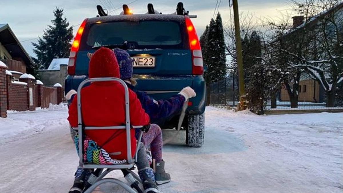 Dwie osoby na sankach ciągniętych przez samoch&oacute;d na zimowej drodze.
