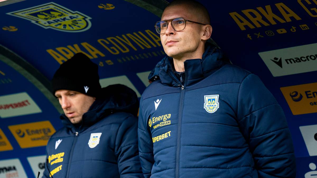 Dw&oacute;ch trener&oacute;w w niebieskich kurtkach sportowych na tle stadionu, jeden w okularach, drugi w czapce.