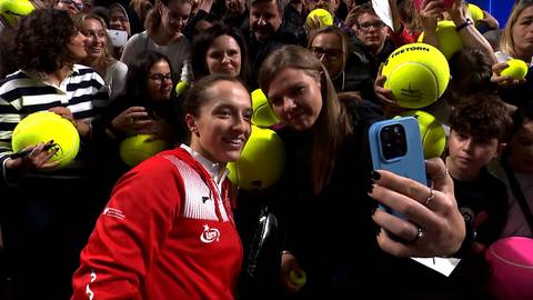 Iga Świątek pozuje do zdjęcia z fanką, która trzyma telefon komórkowy. W tle widać tłum kibiców, z których niektórzy mają piłki tenisowe.