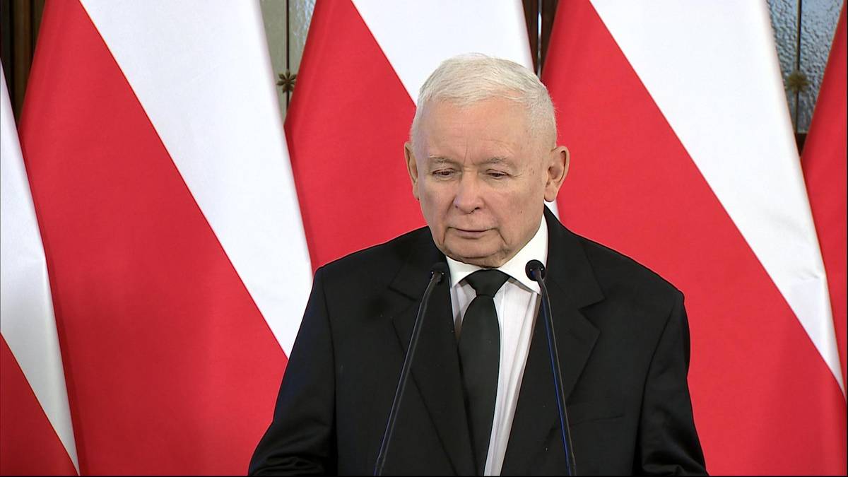 Jarosław Kaczyński w garniturze i krawacie stoi przed mikrofonami na tle flag Polski.