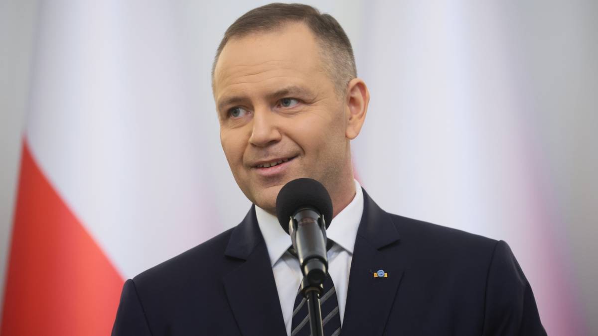 Mężczyzna w garniturze przemawia do mikrofonu, na tle flagi Polski.