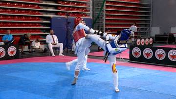 Dw&oacute;ch zawodnik&oacute;w taekwondo w kaskach i ochraniaczach podczas walki na niebiesko-r&oacute;żowej macie w hali sportowej.