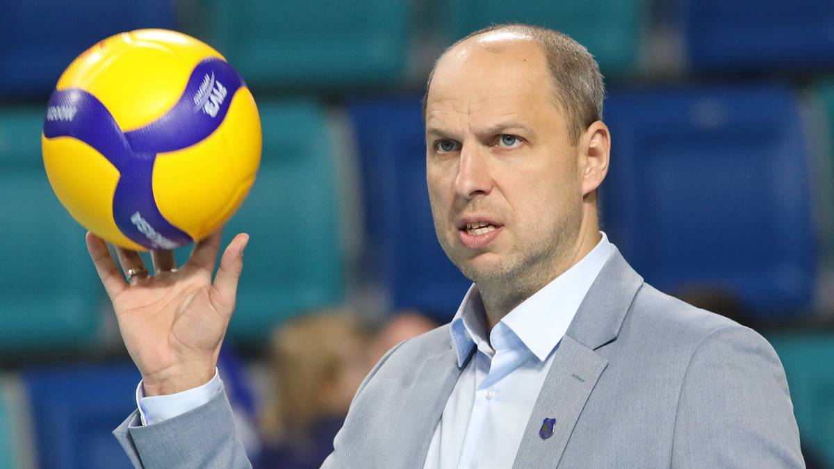 Mężczyzna w szarym garniturze trzyma żółto-niebieską piłkę do siatkówki w prawej dłoni.