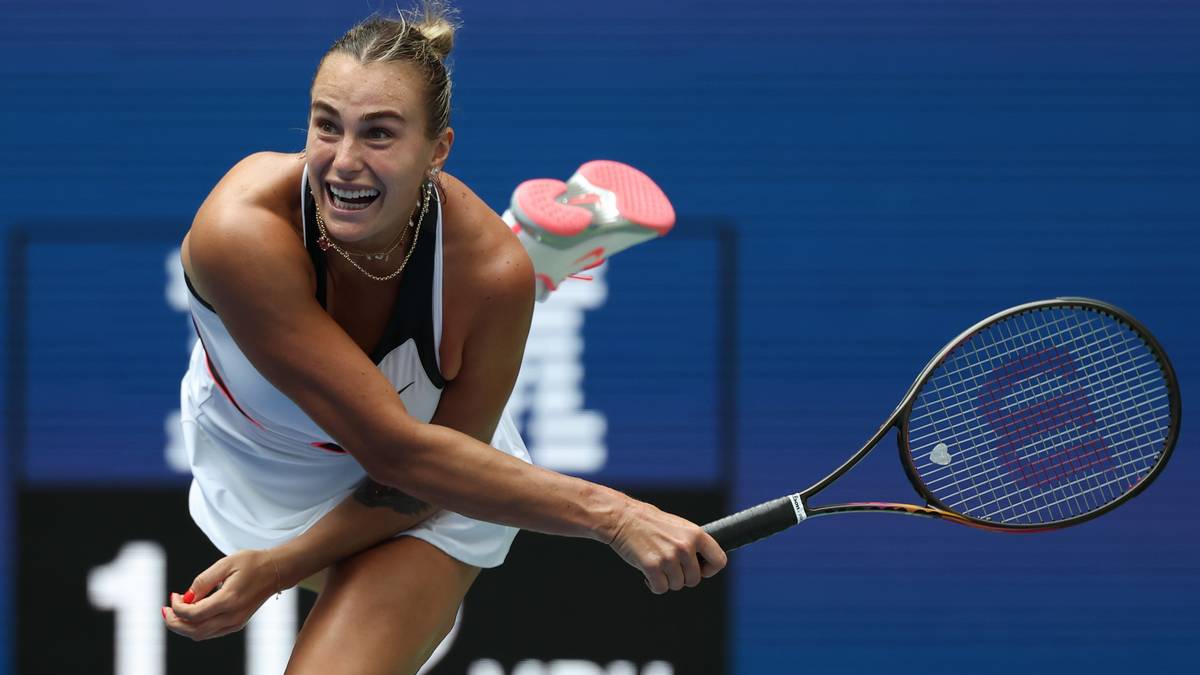Aryna Sabalenka uśmiecha się, wykonując serwis podczas meczu tenisowego. Ma na sobie biały str&oacute;j sportowy i trzyma rakietę tenisową.