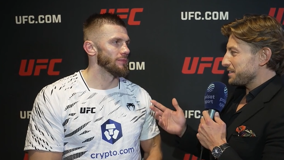 Dwóch mężczyzn rozmawia na tle logo UFC. Jeden z nich, ubrany w białą koszulkę z logo UFC i "crypto.com", ma krótką fryzurę i zarost. Drugi mężczyzna, ubrany w czarny garnitur, trzyma mikrofon i zadaje pytanie.